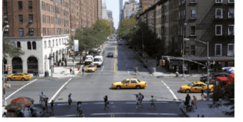 A wide New York street seen from above: yellow taxis and cars at an intersection, people crossing on the crosswalks, apartment blocks and shops on both corners, and tall buildings in the distance.